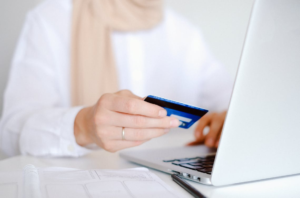  A person in a scarf holding a credit card next to a laptop