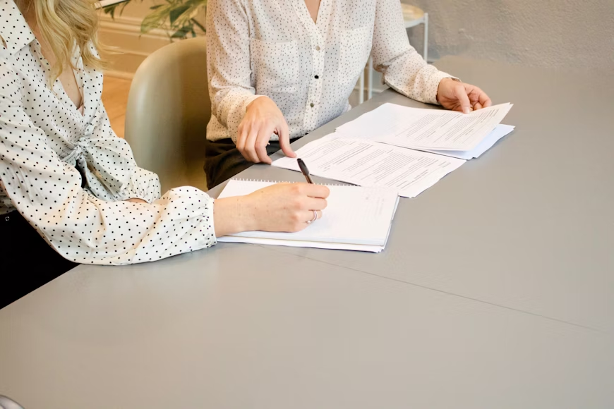 a woman sitting next to another woman, going through papers and signing documents