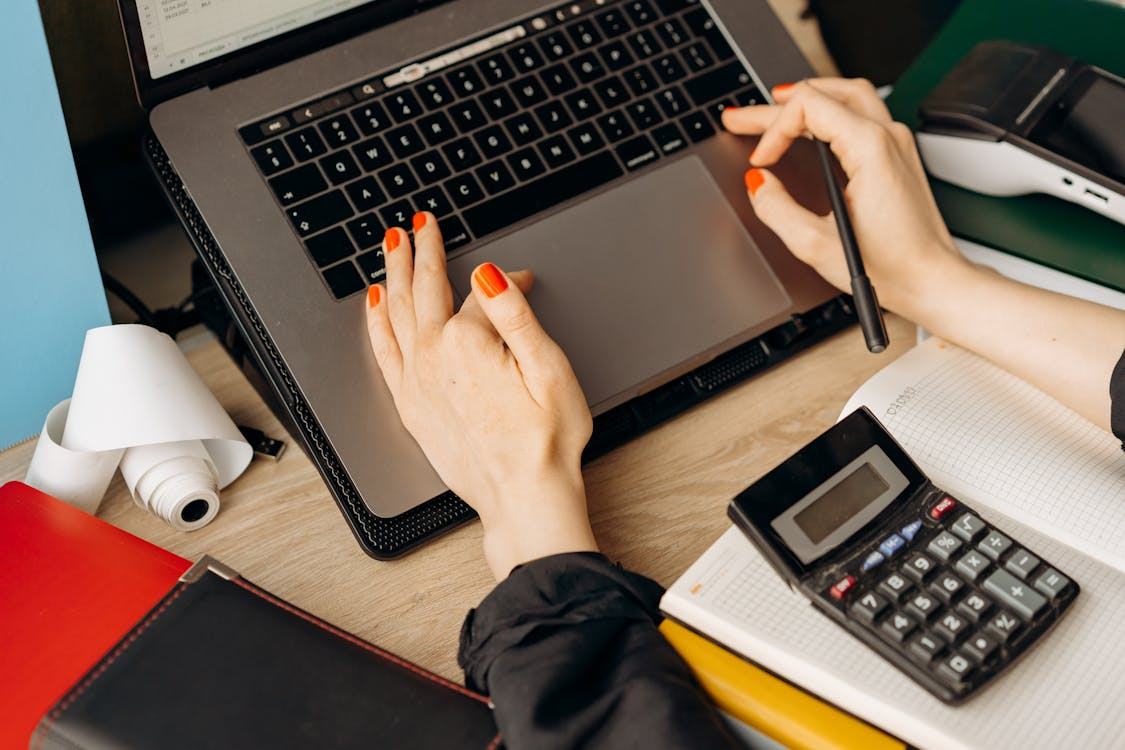 a woman wearing red nail polish using a laptop while holding a pen next to a calculator and a notebook